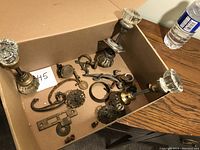 Four vintage glass double-sided door knobs with brass bases alongside various aged metal hardware including handles, plates, and hooks, arranged in a box.