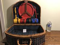 Open wicker picnic basket showing plaid interior and secured accessories—plastic cups, flatware in colorful holders, and a sectioned plate.