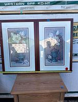 Two framed floral paintings side by side on a table with a tape measure showing size.