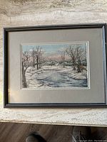 Framed cross stitch artwork showing detailed winter landscape with snow, trees, and river on textured fabric inside a black frame with grey matting.
