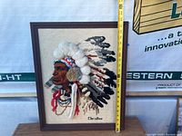Framed cross stitch artwork showing a Native American profile with a 3D headdress and measuring tape indicating height approx. 22 inches.