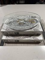 Two oval-shaped clear glass dishes stacked on their boxes, showing patterned cut glass design and scalloped edges.