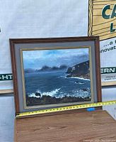 Framed seascape painting resting on a surface showing ocean waves, rocky cliffs and cloudy sky. Signature visible.