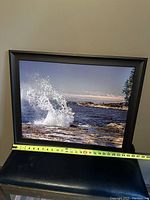 Framed photograph displaying a coastal wave scene with ocean and rocky shoreline, with a tape measure showing width of about 25 inches.