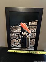Photo of the framed photographic print showing a man with an orange umbrella next to a bicycle and trash can, set against a brick wall, with a measuring tape showing width.