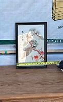 Silk artwork in black frame showing a bird on branch with flowers and calligraphy, placed on table with tape measure showing size.