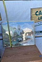 Framed print showing landscape with palm trees, animals, and rustic structure. Roughly 22 inches tall as measured by tape.