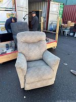 Photo of a single light gray-beige fabric recliner chair with padded armrests and headrest on asphalt surface.