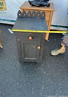 Front and top view of a small rustic cabinet, showing scalloped back edge on top, one door with a wood knob, one drawer with a matching knob, and black paint finish.