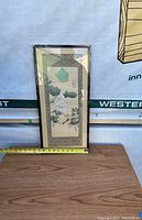 Framed silk screen print placed upright on wooden surface with a measuring tape showing height about 23 inches and width about 11 inches.