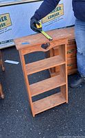 Wooden shelf unit shown upright with a tape measure indicating width of approximately 18 inches and depth, placed outdoors on asphalt ground.