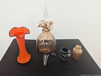 Four items on black surface: clear and amber Murano decanter with stopper, bright orange Tango vase, small cobalt blue jug with stripes, and speckled small vase