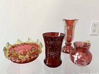 Side angled view of the four cranberry and vaseline art glass pieces including two vases, a pitcher, and a ruffled bowl showing color and decoration.