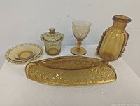 Front view of five amber glass pieces including the canoe bowl, vase, goblet, covered jar on plate, and small plate.