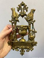 Front view of the antique brass wall hanging match strike showing two birds, a central urn, and ornamental top and base.