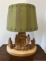 Front view of the carved wood table lamp with green slat-shade and four figures around fireplace base.