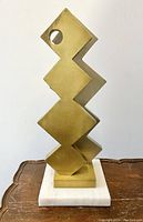 Front view of the gold-toned Brutalist sheet metal sculpture on a white marble base, showing geometric cutout design.