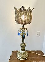 Front view of the lamp illuminated showing the flower-form pierced metal shade, aqua glass prisms, ornate base, and overall lamp height.