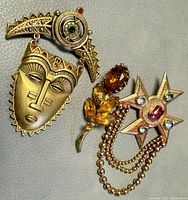 Three vintage costume brooches displayed together, including the AVON tribal mask brooch, a floral design brooch with amber-colored rhinestones, and a star-shaped brooch with rhinestones and gold-tone chains.