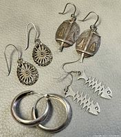 Four pairs of silver earrings shown with varying designs: teardrop filigree, oval with sailboats, fishbone dangle, and smooth hoops.