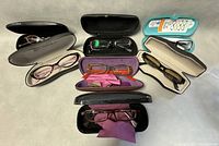 Seven pairs of eyeglasses with their respective hard and soft cases, arranged on a flat surface.