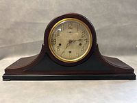 Front view of the clock showing the round clock face, wooden case with curved design, and black base.