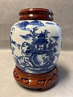 Porcelain ginger jar on wooden carved base, showing detailed blue landscape scenes with pagoda and trees