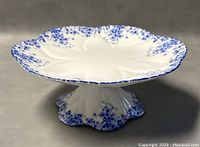 Angled front view of full pedestal cake plate showing overall shape, scalloped rim and blue floral spatter