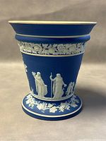 Front view of Wedgwood footed planter showing white classical figures with staffs on blue jasperware background, floral relief border on upper section and base.