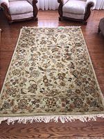 Full view of rectangular wool area rug with floral patterns and fringed edges placed on wooden floor between two chairs.