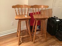 Two wooden bar stools with swivel seats, spindle backrests, and crossbar footrests, in light oak finish.