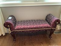 Full view of a wood framed upholstered bench seat with burgundy fabric featuring gold scroll pattern, cylindrical arm rests, and tassel decorations.