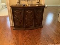 Full view of wood cabinet with three doors closed, showing ornate raised panel designs on the doors and decorative round hardware knobs, standing on hardwood floor.