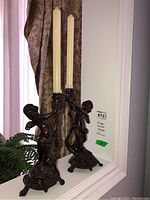 Pair of carved angel candle holders with taper candles inserted, shown on white display ledge near window curtain and greenery.