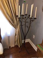 Full view of tall metal candle stand with seven white taper candles, positioned near window and drapes.