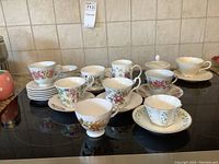 Wide view of various fine china teacups and saucers arranged on a surface. Shows multiple floral pattern china pieces and a white sugar bowl with lid.