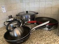 Photo showing multiple cookware items on a kitchen counter including a tea kettle, several pots with lids, and frying pans, showcasing the brands and condition.