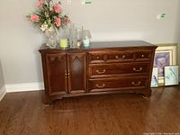 Front view photo of dresser with items on top and paintings leaning against wall beside it.