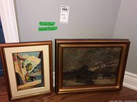 Photo showing both framed paintings side by side on the floor against wall. Larger dark nautical scene to right, smaller bright coastal scene to left.