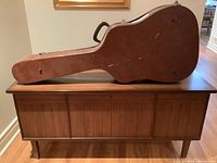 Closed brown wooden guitar case resting on a wood cabinet, viewed from side showing handle and latches.