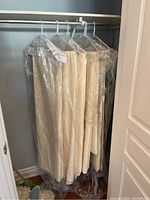 Photograph of five large cream or off-white linens hanging on plastic hangers protected by plastic garment covers in a closet.