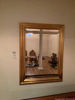 Mirror hanging on wall reflecting room with boxes and white column. Gold-tone frame with beveled edges visible.