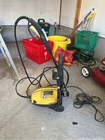 Yellow Karcher power washer unit with attached spray gun, hose, and power cable on a concrete floor inside a garage with various other items in background.