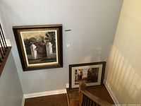 Photo showing two framed prints hanging on a wall by a staircase, one larger above the other leaning against the wall.
