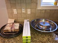 Overview photo showing stacked serving trays, boxed ceramic dishes, and stainless steel bowl with covered dish on a kitchen counter.