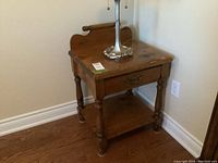 Front angled view of vintage wooden side table with drawer, lower shelf, and decorative backboard with curled edges. Shows wood grain and scratches on surface.