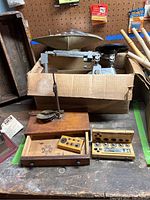 Photo showing hanging scale on wooden box with drawer, metal baker's scale in cardboard box and two wooden boxes with weights.