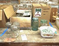 Overview of two open cardboard boxes with various glass jars inside, several jars outside the box include Atlas and Ball glass jars, and a white ceramic casserole dish with floral design.