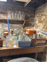 Wide shot of various clear glass jars and bottles of different sizes on wooden table with brick wall background. Some bottles have lids, others open.