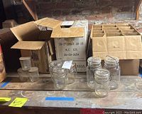 Several clear glass canning jars and smaller jelly jars displayed on a shelf with cardboard boxes behind them.
